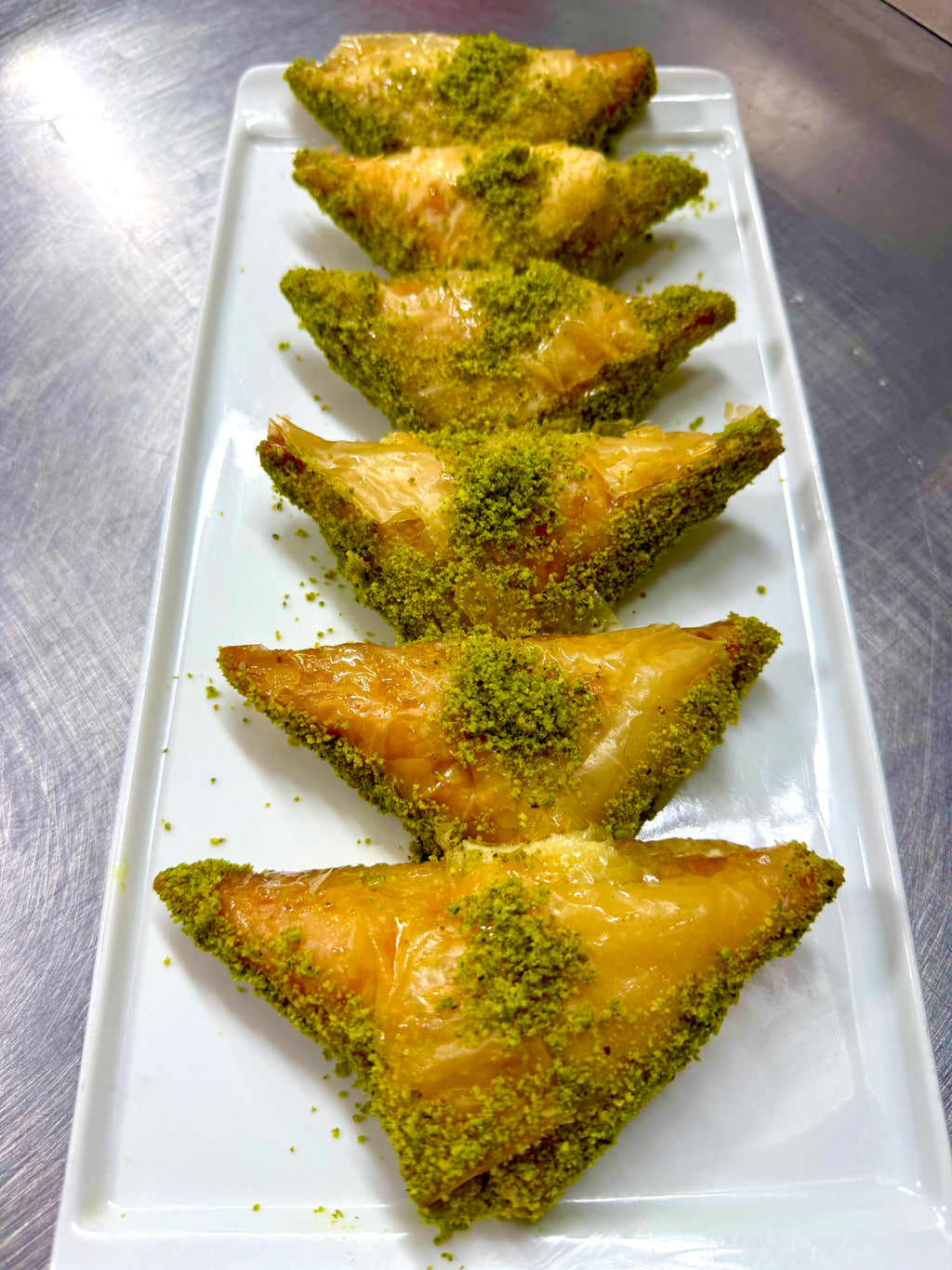 Triangular pieces of sobiyet filled with homemade cream topped with green pistachio on a white rectangular plate.