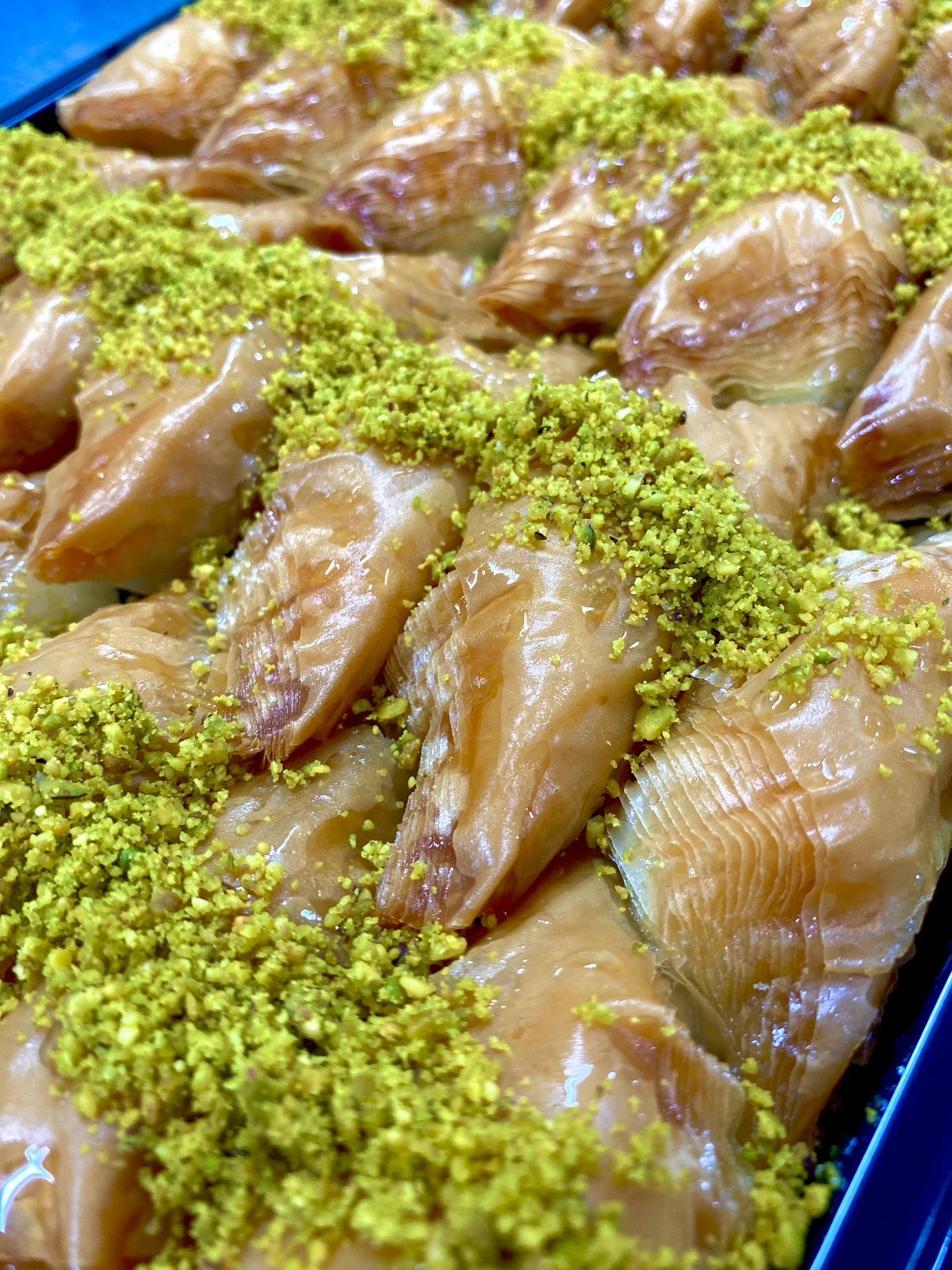 Close-up of golden folded Sobiyet baklava triangles generously topped with bright green ground pistachios and filled with a delicious cream.
